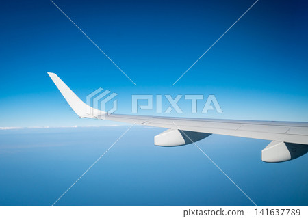 Airplane wing above blue sky and distant clouds viewed from passenger window during an international commercial airline flight. Aviation, travel, transportation and global tourism. Aviation industry, Airplane wing above blue sky and distant clouds viewed from passenger window during an international commercial airline flight. Aviation, travel, transportation and global tourism. Aviation industry, 141637789