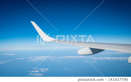 Commercial airplane wing seen from passenger window while flying above clouds under clear blue sky. Modern aviation, international flight, travel experience and tourism concept. Global tourism. 141637790