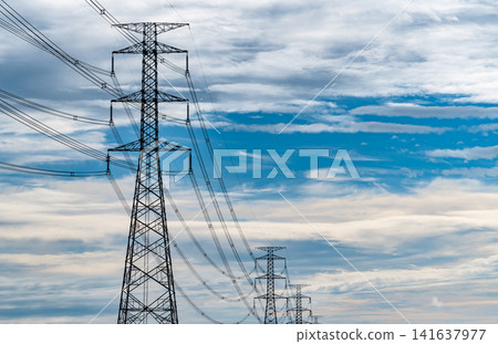 High voltage electricity transmission towers and power lines beneath a blue cloudy sky. Modern electrical infrastructure, utility grid reliability, energy distribution and sustainable power networks. High voltage electricity transmission towers and power lines beneath a blue cloudy sky. Modern electrical infrastructure, utility grid reliability, energy distribution and sustainable power networks. 141637977