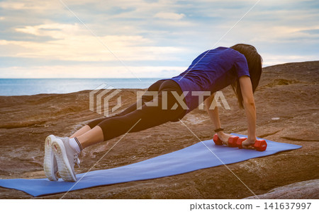 Asian woman performing dumbbell plank exercise on a yoga mat beside a tropical beach at sunrise, combining outdoor strength training, core workout, functional fitness and healthy active lifestyle. Asian woman performing dumbbell plank exercise on a yoga mat beside a tropical beach at sunrise, combining outdoor strength training, core workout, functional fitness and healthy active lifestyle. 141637997