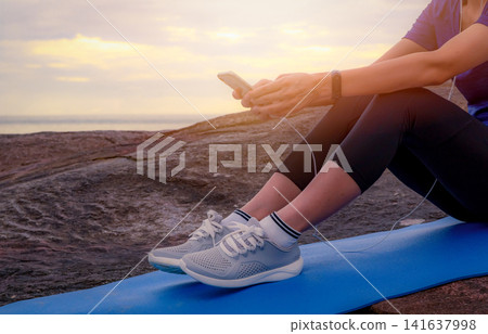 Asian woman resting after a morning beach workout while checking smartphone and fitness tracker at sunrise. Wearable technology, wellness monitoring, active lifestyle and tropical coastal scenery. 141637998