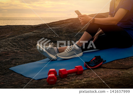Asian woman relaxing after outdoor strength workout while checking her smartphone beside the tropical sea at sunrise. Dumbbells, wearable fitness tracker, recovery routine and healthy active lifestyle Asian woman relaxing after outdoor strength workout while checking her smartphone beside the tropical sea at sunrise. Dumbbells, wearable fitness tracker, recovery routine and healthy active lifestyle 141637999