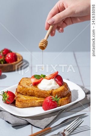 The hand holds a stick that drips honey on to brioche toasts with strawberries and whipped cream The hand holds a stick that drips honey on to brioche toasts with strawberries and whipped cream 141638098