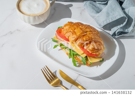 A golden croissant sandwich with ham, cheese, tomato, and arugula on a marble background 141638132