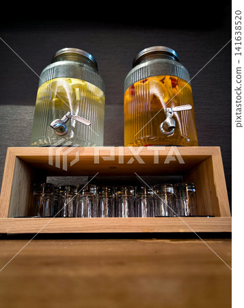 Two glass beverage dispensers with fruit infused water on wooden stand above stacked drinking glasses in modern catering setup, refreshing infused drinks in stylish conference buffet background Two glass beverage dispensers with fruit infused water on wooden stand above stacked drinking glasses in modern catering setup, refreshing infused drinks in stylish conference buffet background 141638520