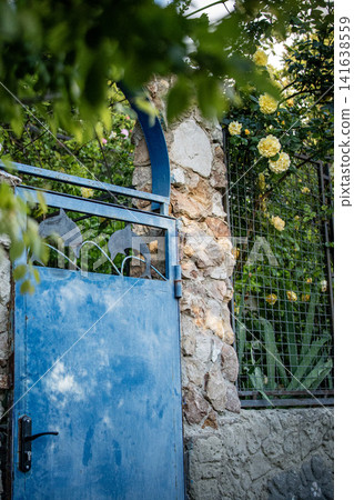 Blue old rustic garden gate with stone wall and rose bush in rural village - Eastern Europe Blue old rustic garden gate with stone wall and rose bush in rural village - Eastern Europe 141638559