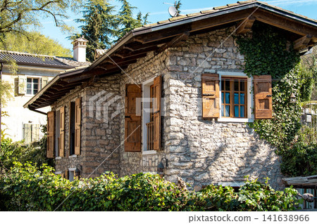 Rustic Stone House With Wooden Shutters: Traditional Alpine Architecture In Northern Italy Surrounded By Greenery 141638966