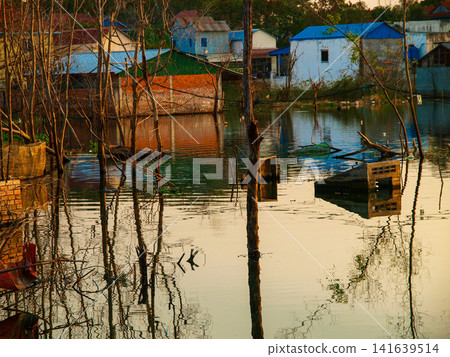 Traditional stilt houses and structures reflected in calm water during golden hour in a rural village near Phnom Penh, Cambodia Traditional stilt houses and structures reflected in calm water during golden hour in a rural village near Phnom Penh, Cambodia 141639514