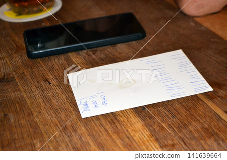 Traditional bill on a piece of paper in a Prague pub. Wooden table scene with bill, receipt phone and beer ring stains in prague pub for background and lifestyle poster design 141639664