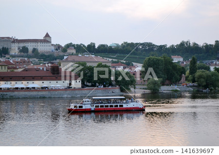 Red and white sightseeing cruise boat on the vltava river in Prague with riverside architecture and green embankment for travel background banner and poster design Red and white sightseeing cruise boat on the vltava river in Prague with riverside architecture and green embankment for travel background banner and poster design 141639697