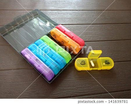 A black pill organizer with seven multicolored plastic containers. One of them is open A black pill organizer with seven multicolored plastic containers. One of them is open 141639781