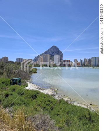 Scenic view of salt lake Las Salinas in Calpe with Penon de Ifach mountain and green vegetation under blue sky Scenic view of salt lake Las Salinas in Calpe with Penon de Ifach mountain and green vegetation under blue sky 141640330