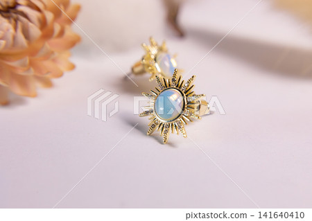 Elegant gold ring with a blue gemstone displayed on a soft pink surface beside a dried flower, creating a delicate jewelry composition with romantic styling and luxury aesthetic Elegant gold ring with a blue gemstone displayed on a soft pink surface beside a dried flower, creating a delicate jewelry composition with romantic styling and luxury aesthetic 141640410