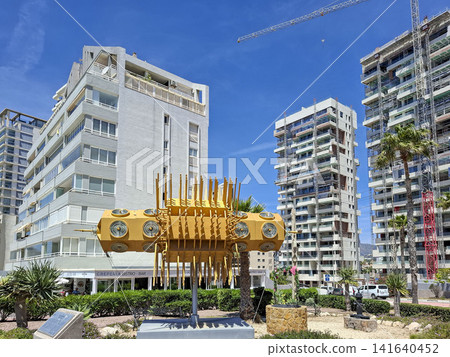 Calpe, Spain - May 27, 2026.Ornate gold kinetic sculpture in city park with modern buildings and construction cranes in the background Calpe, Spain - May 27, 2026.Ornate gold kinetic sculpture in city park with modern buildings and construction cranes in the background 141640452