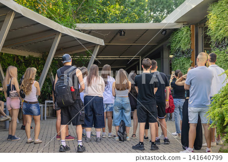 Visitors Gather at Outdoor Public Event. Group of young people and adults stand together, outdoor public event in garden venue Visitors Gather at Outdoor Public Event. Group of young people and adults stand together, outdoor public event in garden venue 141640799