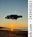 Silhouette of Flying Drone Over Beach at Sunset by the Sea 141641623