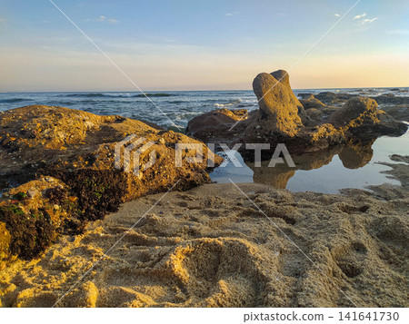 Golden sunset light illuminates a rocky shoreline with a peaceful tide pool reflecting jagged stones. A serene coastal landscape ideal for travel, nature, and wellness marketing. Golden sunset light illuminates a rocky shoreline with a peaceful tide pool reflecting jagged stones. A serene coastal landscape ideal for travel, nature, and wellness marketing. 141641730