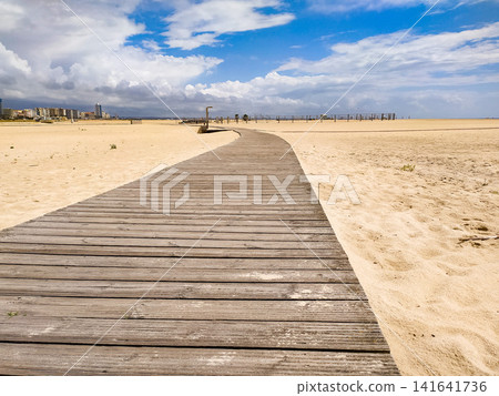 This winding wooden boardwalk path leads across a vast sandy beach under a beautiful, dramatic blue sky, evoking a sense of a peaceful summer journey and solitude with absolutely no people. This winding wooden boardwalk path leads across a vast sandy beach under a beautiful, dramatic blue sky, evoking a sense of a peaceful summer journey and solitude with absolutely no people. 141641736