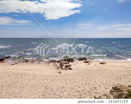 Serene beach landscape with rocky shore, footprints on sand, and vast blue ocean under a bright cloudy sky. Nobody. Evoking tranquility and peace, perfect for travel concepts. Serene beach landscape with rocky shore, footprints on sand, and vast blue ocean under a bright cloudy sky. Nobody. Evoking tranquility and peace, perfect for travel concepts. 141641739