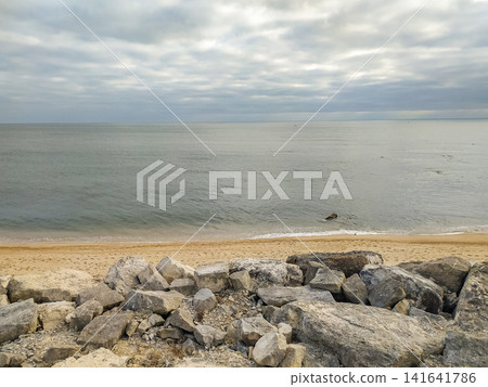 A serene coastal landscape with no people featuring rugged rocks in the foreground, golden sand, and a vast, calm ocean under a moody, overcast sky. It evokes peace, solitude, and resilience. A serene coastal landscape with no people featuring rugged rocks in the foreground, golden sand, and a vast, calm ocean under a moody, overcast sky. It evokes peace, solitude, and resilience. 141641786