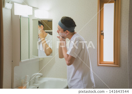 A man in his 40s, seen from behind, washing his face with foaming soap in front of a bathroom mirror. A man in his 40s, seen from behind, washing his face with foaming soap in front of a bathroom mirror. 141643544