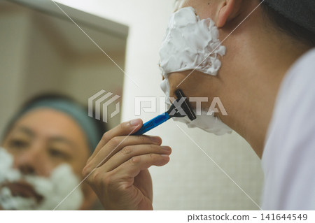 A man in his 40s shaves his chin against the grain with a razor in the bathroom. A man in his 40s shaves his chin against the grain with a razor in the bathroom. 141644549