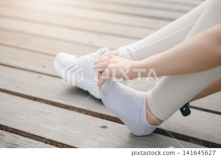 Woman stretching foot after running with muscle pain and leg injury, female athlete sits on wooden boardwalk holding painful foot in white sock during workout recovery outdoors Woman stretching foot after running with muscle pain and leg injury, female athlete sits on wooden boardwalk holding painful foot in white sock during workout recovery outdoors 141645627
