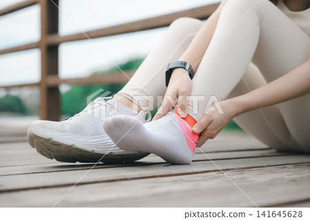Injured runner woman experiences sudden ankle injury pain, sitting on outdoor wooden floor for rest and physical pressure relief after fitness running exercise to ease joint inflammation 141645628