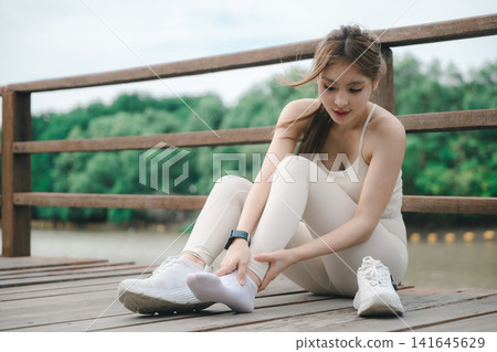 Woman runner athlete holding ankle, suffering muscle tendon pain, feeling intense sport injury discomfort while sitting on wooden bridge outdoor, needing medical treatment Woman runner athlete holding ankle, suffering muscle tendon pain, feeling intense sport injury discomfort while sitting on wooden bridge outdoor, needing medical treatment 141645629