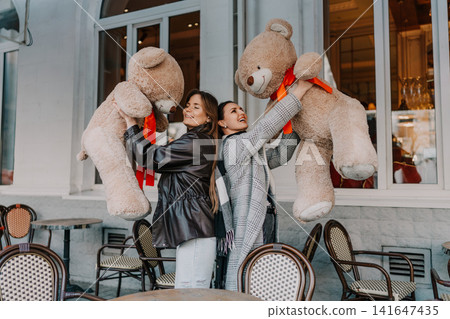 Friends women bears laughing and lifting giant plush toys while standing outside an urban cafe on a sunny autumn day Friends women bears laughing and lifting giant plush toys while standing outside an urban cafe on a sunny autumn day 141647435