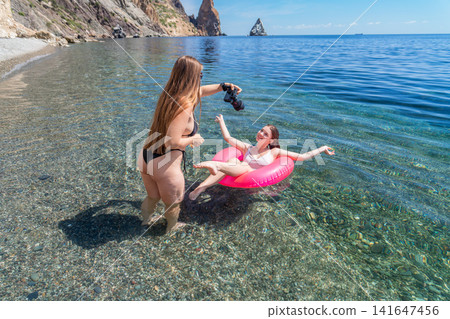 Woman Beach Photographer taking a photo of her friend on a pink inflatable ring in the clear turquoise sea during a sunny summer vacation Woman Beach Photographer taking a photo of her friend on a pink inflatable ring in the clear turquoise sea during a sunny summer vacation 141647456