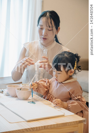 一位家長和孩子在房間裡玩耍；一個女孩正在用顏料畫畫。 141648504