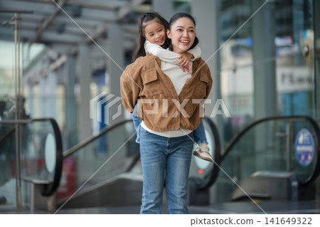 happy mother giving daughter piggyback ride near escalator in shopping mall or department store 141649322