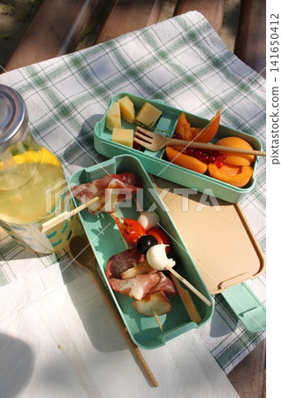 Colorful picnic food arranged in green containers with skewers, accompanied by a glass of lemonade on a checkered tablecloth outdoors in a sunny setting Colorful picnic food arranged in green containers with skewers, accompanied by a glass of lemonade on a checkered tablecloth outdoors in a sunny setting 141650412