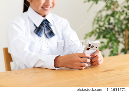 A middle school student in uniform is looking at her smartphone at the living room table. A middle school student in uniform is looking at her smartphone at the living room table. 141651771
