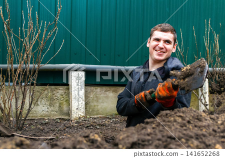 A young man smiling while gardening, digging the soil with a shovel in the yard near green bushes and a fence, illustrating gardening, country life, and agriculture A young man smiling while gardening, digging the soil with a shovel in the yard near green bushes and a fence, illustrating gardening, country life, and agriculture 141652268