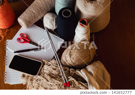 Crafting and productivity concept with various yarn balls. Knitting needles. A notepad. Pen. Scissors. And a smartphone on a wooden table Crafting and productivity concept with various yarn balls. Knitting needles. A notepad. Pen. Scissors. And a smartphone on a wooden table 141652777
