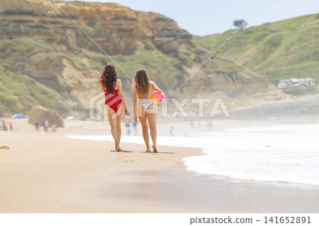 Two women in swimsuits walking along a sandy beach, one in red and the other in white, with cliffs and ocean waves in the background on a sunny day Two women in swimsuits walking along a sandy beach, one in red and the other in white, with cliffs and ocean waves in the background on a sunny day 141652891