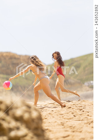 Two young women playing with a colorful beach ball on sandy shore, wearing swimsuits, with green hills and clear sky in the background Two young women playing with a colorful beach ball on sandy shore, wearing swimsuits, with green hills and clear sky in the background 141652892
