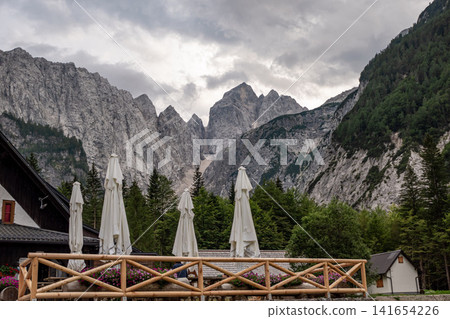View from Tamar House in Slovenia, alpine mountain lodge with wooden terrace, green meadow, pine forest and dramatic Julian Alps peaks under cloudy sky View from Tamar House in Slovenia, alpine mountain lodge with wooden terrace, green meadow, pine forest and dramatic Julian Alps peaks under cloudy sky 141654226