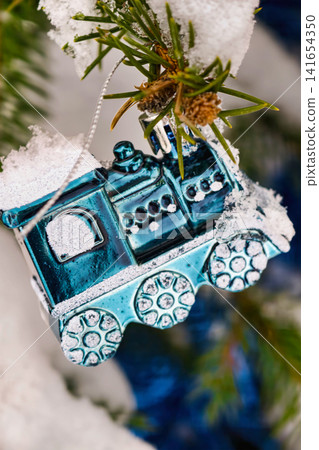 A blue train ornament hangs from a green Christmas tree covered in snow. Bright lights and other decorations surround the tree, adding to the holiday spirit A blue train ornament hangs from a green Christmas tree covered in snow. Bright lights and other decorations surround the tree, adding to the holiday spirit 141654350