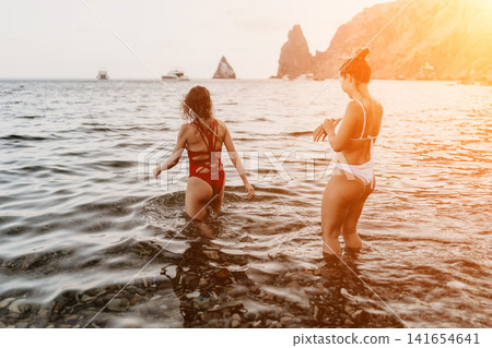 Women beach ocean, two friends walking into the sea from a rocky shore during sunset. Women beach ocean, two friends walking into the sea from a rocky shore during sunset. 141654641
