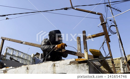 Manual laborer in protective mask welds iron detail at construction. Professional young repairman with welding machine working at the site. Male welder operating with metal on house. Close up Manual laborer in protective mask welds iron detail at construction. Professional young repairman with welding machine working at the site. Male welder operating with metal on house. Close up 141654746