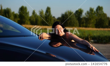 Beautiful smiling girl bought new electrical vehicle and hugging hood auto. Happy young woman enjoying modern electric automobile. Concept of buy or rent car. Slow motion Beautiful smiling girl bought new electrical vehicle and hugging hood auto. Happy young woman enjoying modern electric automobile. Concept of buy or rent car. Slow motion 141654776