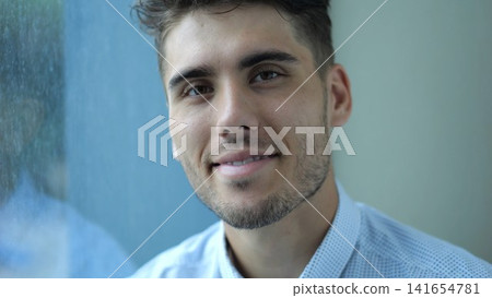 Portrait of happy businessman sitting near window in modern hotel room or office. Relaxed young manager looks into camera with smile showing relief, confidence and success in professional lifestyle Portrait of happy businessman sitting near window in modern hotel room or office. Relaxed young manager looks into camera with smile showing relief, confidence and success in professional lifestyle 141654781