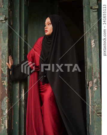 Portrait of a beautiful young Muslim woman wearing an elegant black abaya and red dress posing confidently in a rustic industrial warehouse, modern modest fashion concept. Portrait of a beautiful young Muslim woman wearing an elegant black abaya and red dress posing confidently in a rustic industrial warehouse, modern modest fashion concept. 141655113