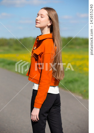 Young woman enjoys sunny day outdoors while wearing stylish orange jacket and black pants Young woman enjoys sunny day outdoors while wearing stylish orange jacket and black pants 141655240