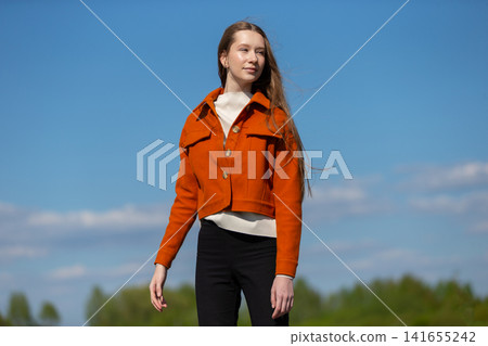 Young woman in stylish orange jacket and black pants stands confidently outdoors under a clear blue sky Young woman in stylish orange jacket and black pants stands confidently outdoors under a clear blue sky 141655242