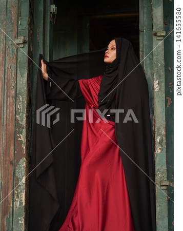 Portrait of a beautiful young Muslim woman wearing an elegant black abaya and red dress posing confidently in a rustic industrial warehouse, modern modest fashion concept. Portrait of a beautiful young Muslim woman wearing an elegant black abaya and red dress posing confidently in a rustic industrial warehouse, modern modest fashion concept. 141655580