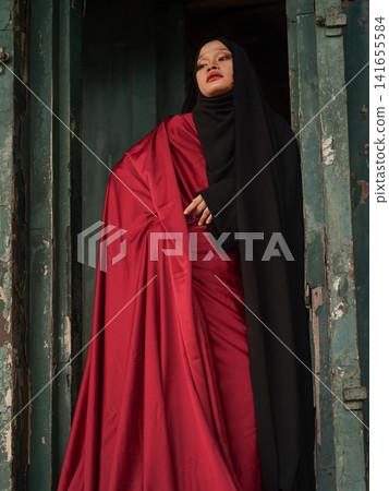 Portrait of a beautiful young Muslim woman wearing an elegant black abaya and red dress posing confidently in a rustic industrial warehouse, modern modest fashion concept. Portrait of a beautiful young Muslim woman wearing an elegant black abaya and red dress posing confidently in a rustic industrial warehouse, modern modest fashion concept. 141655584